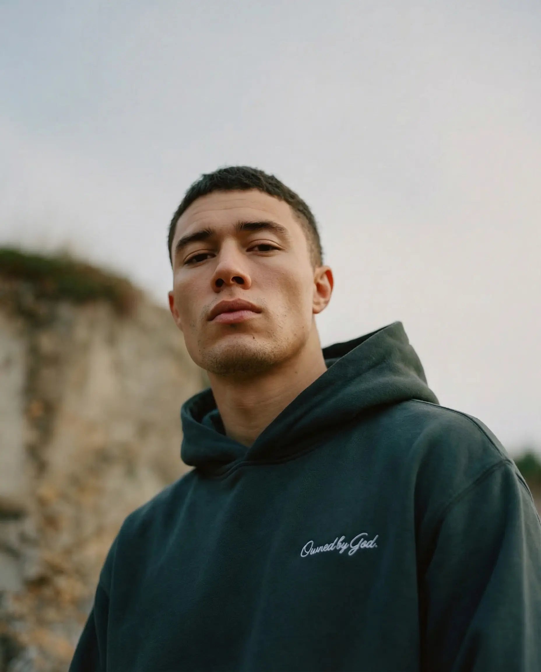 Male model wearing CORE Monogram Hoodie in Pine Green by Owned by God — relaxed oversized fit, tonal embroidery on chest and cuff, outdoors.